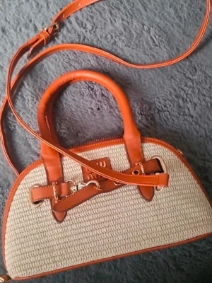 Beige och brun handväska  - Snygg handväska i beige flätad textil med bruna läderdetaljer och axelrem. Väskan har guldfärgade metalldetaljer och dubbla handtag. Perfekt accessoar för dig som gillar en trendig och stilren look.