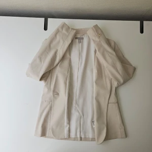 Beige kavaj från H&M, storlek 42 - Stilren beige kavaj från H&M i storlek 42. Klassisk passform med två fickor framtill och ett knappslå. Tillverkad i ett mjukt tyg med helfodrad insida för extra komfort. Perfekt för att lyfta din outfit med en clean och modern look.