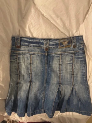 Blå jeanskjol med fickor och detaljer - Säljer en blå jeanskjol med markerade sömmar, breda hällor och snygga fickor både fram och bak. Kjolen har plisserade detaljer nedtill och en liten broderad logga framtill. Perfekt för dig som gillar en avslappnad men trendig stil.