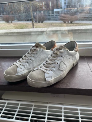  Philippe Model skor  - Snygga vita sneakers från Philippe Model med beige mockadetaljer på tå och plös samt bronsfärgad häl. Skorna har klassisk låg profil, vita skosnören och en diskret logotyp på sidan. Perfekta för dig som gillar stilrena och trendiga sneakers.