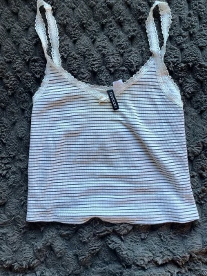 Randig linnetopp med spets från Divided - Söt vit och svart randig linnetopp från Divided med spetsdetaljer vid axelband och urringning. Toppen har en liten rosett framtill och är croppad med smala axelband. Perfekt för en avslappnad och trendig look.