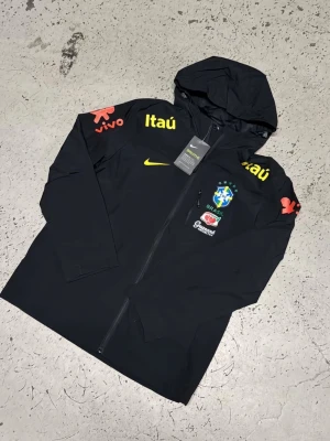 Svart Brasil Nike träningsjacka - Svart träningsjacka från Nike med huva och dragkedja. Jackan har Brasilien fotbollslandslagets logga på bröstet samt sponsorloggor i gult, vitt och rött. Tillverkad i lätt funktionsmaterial, perfekt för sport och träning.