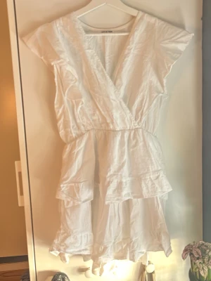 Vit omlottklänning med volanger - Säljer en vit kortklänning från Meet Me There med omlott-design och volangdetaljer. Klänningen har korta ärmar och är tillverkad i ett lätt bomullsmaterial. 