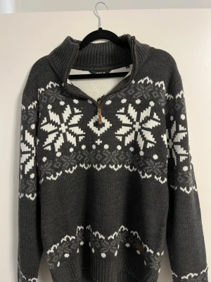 Grå stickad tröja med snöflingor - Mysig grå stickad tröja från !Solid med vita snöflingor och klassiskt vintermönster. Tröjan har halv dragkedja, hög krage och ribbade muddar. 