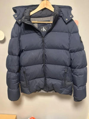Calvin Klein jacka - Snygg mörkblå pufferjacka från Calvin Klein Jeans med huva och dragkedja framtill. Jackan har två framfickor med dragkedja och en patch med logga på ärmen. Perfekt för kalla vinterdagar och riktigt skön att bära. Har inte använt denna mycket eftersom den är lite för stor för mig fick i present av min storebror då den inte passar honom!