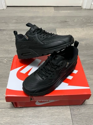 Nike Air Max 90 svart sneakers - Snygga helsvarta Nike Air Max 90 sneakers med klassisk siluett. Ovandelen är i en mix av läder och mesh för både stil och komfort. Tydlig Air Max-dämpning i sulan och diskreta detaljer som ger en clean look. Perfekta för dig som gillar streetwear och sportig stil.