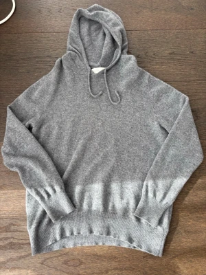 Grå cashmere hoodie från Sayless - Mjuk och stilren grå hoodie från Sayless i lyxig cashmere. Klassisk modell med huva och dragsko, ribbade muddar vid ärmslut och nederkant. Perfekt för dig som gillar en enkel men exklusiv look.