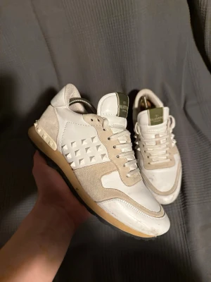 Valentino rockrunner  - Snygga vita sneakers från Valentino med beige mockadetaljer och ikoniska pyramidnitar på sidorna och hälen. Skorna har en grön logga på plösen, vita snören och en beige sula. Perfekta för dig som vill ha en unik och trendig look.