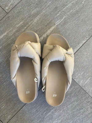 Beige sandaler med knut från H&M - Säljer ett par snygga beige sandaler från H&M i storlek 37. De har en bred rem med trendig knutdetalj och är gjorda i ett mjukt, vadderat material som känns skönt mot foten. Använda få gånger