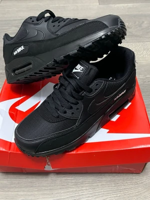 Nike Air Max 90 helsvarta sneakers - Snygga helsvarta Nike Air Max 90 sneakers med klassisk siluett. Skorna har ovandel i textil och läder, synlig Air-enhet i sulan och vita Air Max-loggor på sidan. Perfekta för dig som gillar stilrena och tidlösa sneakers.