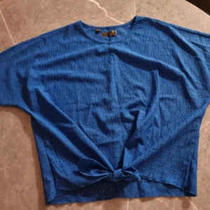 Blå kortärmad topp med knyt - Blå topp i bomull med korta ärmar och rund hals. Toppen har ett fint hålmönster över hela tyget och en knytning framtill som ger en avslappnad look. Perfekt för dig som gillar färg och detaljer.