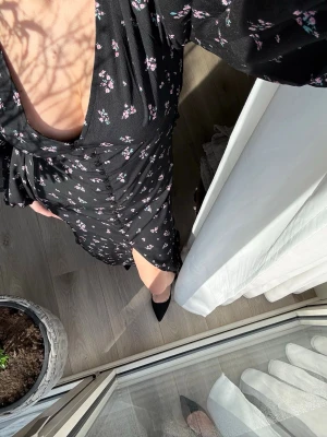 Svart blommig långklänning med v-ringning - Svart långklänning med små rosa och vita blommor, djup v-ringning och långa ärmar. Klänningen har en markerad midja och lätt slits framtill. 