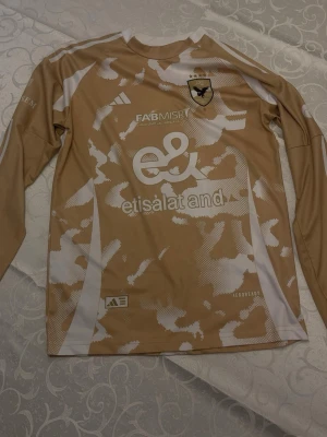 Beige långärmad fotbollströja Adidas - Snygg beige långärmad fotbollströja från Adidas med vitt kamouflagemönster och flera tryckta loggor, bland annat etisalat, FAB MISR och Shell Helix. Tröjan har nummer 22 på ryggen och är tillverkad i ett lätt och ventilerande material.