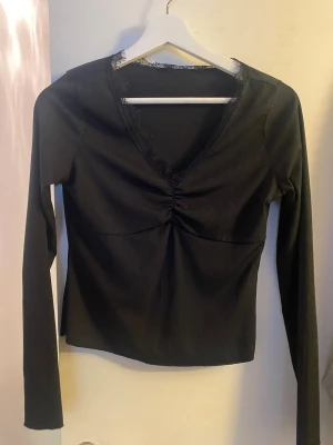 Svart långärmad topp med spets - Snygg svart långärmad topp med ribbad struktur och v-ringning dekorerad med spetsdetaljer. Toppen har en rynkad detalj framtill som ger en cool form. Perfekt att styla med jeans eller kjol för en trendig look.