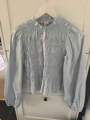 Ljusblå smockad blus med volang - Trendig ljusblå blus från NA-KD med smockad överdel, volangkant nedtill och ballongärmar. Blusen har knappar framtill och en liten ståkrage. Tillverkad i ett lätt bomullsliknande material som ger en luftig känsla.