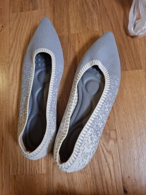 Grå stickade ballerinaskor - Snygga ballerinaskor i stickat material med spetsig tå och gråmelerat mönster. Skorna har en ljusgrå bas med vita detaljer runt öppningen och en bekväm innersula med struktur. Perfekta för dig som gillar minimalistisk och modern stil.