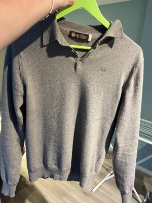 Grå pikétröja i ull från Morris - Snygg grå långärmad pikétröja från Morris med klassisk krage och två knappar vid halsen. Tröjan är tillverkad i mjuk ull och har diskret logga broderad på bröstet. Perfekt för en stilren och avslappnad look.