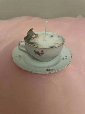 Vanilj doftljus i en vit tekopp med färgglada blommor på   - Det här ljuset är gjort av återanvändbara material. Tekoppen är köpt secondhand och stearinet är från kyrkor som tänkte slänga det. Det här ljuset har en svag doft utav vanilj, så känsliga även kan använda det. Det här ljuset har även torkade blommor på som dekoration. 