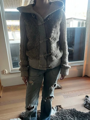 Grå stickad hoodie med fuskpäls - Mysig grå stickad hoodie med fuskpäls framtill och stora träknappar. Tröjan har breda ribbade muddar, luva och en avslappnad passform. Perfekt för lager på lager och en cool streetstil.