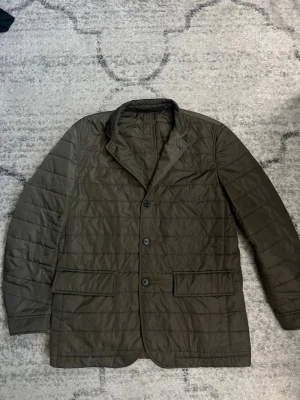 Olivgrön quiltad dunjacka - Stilren olivgrön dunjacka med quiltat mönster och klassisk kavajkrage. Jackan har två stora framfickor och stängs med tre knappar framtill. Perfekt för kalla vinterdagar och ger en snygg, modern look.