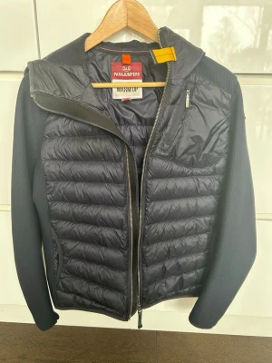 Mörkblå  hybrid jacka från Parajumpers stl YM (14 år) - Snygg mörkblå hybridjacka från Parajumpers med quiltad framsida och stickade ärmar. Jackan har dragkedja framtill, en bröstficka med dragkedja och en gul detalj vid kragen. Perfekt för lager på lager och streetstyle. Sparsamt använd och mycket bra skick. Inga skador. Storleksguide från pjs ligger som sista bild. 