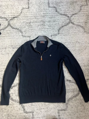 Mörkblå stickad tröja från Morris - Snygg mörkblå stickad tröja från Morris med halv dragkedja och kontrasterande ljusgrå insida på kragen. Tröjan har en liten broderad logga på bröstet och är tillverkad i 100% merinoull. Perfekt till lager på lager och passar till allt.