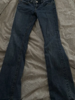 Mörkblå bootcut jeans med fickdetaljer - Snygga mörkblå bootcut jeans med klassisk femficksdesign och dekorativa ficklock med knappar bak. Jeansen har normal midja och är tillverkade i ett mjukt denimtyg som ger en skön passform. Perfekta till sneakers eller boots.