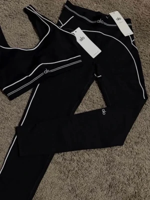 Svarta leggings från Alo med vita detaljer - Snygga svarta leggings från Alo med vita kontrastsömmar längs benen och midjan. Tillverkade i stretchigt material som sitter tight och bekvämt. Perfekta för träning eller chill, med diskret Alo-logga på benet. Matcha gärna med sport-bh i samma stil för en komplett look.