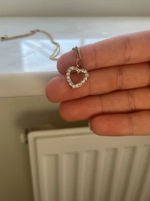 Guldhalsband med hjärthänge och pärlor - Snyggt halsband i guldton med ett hjärtformat hänge dekorerat med små vita pärlor. Kedjan är tunn och elegant, perfekt för att ge en söt touch till din outfit. Hjärtat är öppet i mitten och ger ett fint intryck.