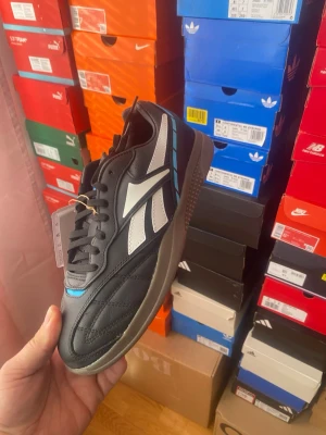 Svarta Reebok sneakers med vita detaljer - Snygga svarta Reebok sneakers med vita och ljusblå detaljer på sidan. Skorna har en klassisk låg siluett, svart ovandel i läder och mocka samt en brun gummisula. Perfekta för dig som gillar retrostil och streetwear.