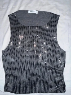 Glittrig svart linnetopp från FB sister - Säljer en glittrig svartfärgad linnetopp från FB sister. Toppen är ärmlös och har paljettdetaljer över hela framsidan samt knytband i ryggen. Perfekt för dig som vill sticka ut med en cool och trendig look.
