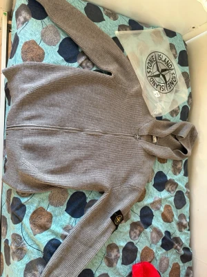 Grå ziptröja från Stone Island - Säljer en grå ribbstickad ziptröja från Stone Island med hög krage och ikonisk patch på ärmen. Tröjan har hel dragkedja framtill och är tillverkad i ett mjukt, bekvämt material. Perfekt för dig som gillar stilrena och exklusiva plagg.