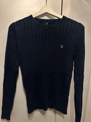 Mörkblå kabelstickad tröja från Gant - Snygg mörkblå kabelstickad tröja från Gant med rund halsringning och diskret logga broderad på bröstet. Tröjan har långa ärmar och ribbade muddar vid ärmslut och nederkant. Perfekt för en stilren och klassisk look.