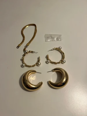 Två par guldiga örhängen + armband - Säljer två par örhängen och ett armband i guldton. Ett par är chunky och halvmåneformade, det andra är ringar med textur och dekorerade med vita pärlor. Perfekta för dig som vill sticka ut med trendiga accessoarer. Örhängena är ej använda.