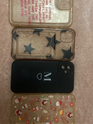 Fyra mobilskal med olika motiv - Säljer fyra unika mobilskal. Ett transparent med vit text, ett transparent med svarta stjärnor och prickar, ett svart med ett vitt M och hjärta, samt ett transparent med julmotiv i rött och vitt. Alla skal är i plast och har olika kreativa designer. Alla är för iPhone 11 och säljer alla tillsammans för 40kr men kan diskutera om man vill köpa någon separat
