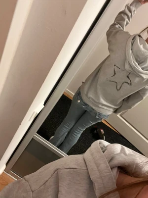 Grå hoodie med stjärna och text - Säljer en ljusgrå hoodie med huva och snörning. På bröstet står det 'Amour' i svart/vitt mönster och på ryggen finns en stor stjärna. Hoodien har en avslappnad passform och är gjord i mjukt sweatshirtmaterial. Perfekt för en chill och casual stil. Finns dock en gul fläck på vänstra akten som tyvärr inte går bort men som knappt syns.