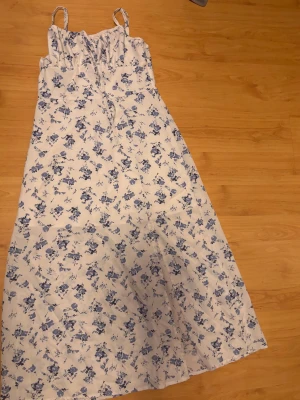 Vit långklänning med blå blommor - Söt långklänning med smala axelband och blått blommönster på vit botten. Klänningen har en feminin siluett och är perfekt för varma dagar. Materialet känns lätt och luftigt, troligen bomull eller polyester. Passar dig som gillar romantisk och somrig stil.