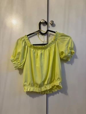 Gul off shoulder topp med puffärm - Trendig gul off shoulder topp med puffiga korta ärmar och resår vid hals, ärmslut och midja. Tillverkad i ett lätt och luftigt tyg som ger en somrig vibe. Perfekt för dig som vill sticka ut med färg och volym i garderoben. Prislapp sitter kvar.