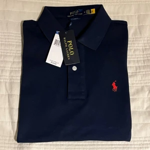 Mörkblå pikétröja från Polo Ralph Lauren - Klassisk mörkblå pikétröja från Polo Ralph Lauren med röd broderad logga på bröstet. Tröjan har krage, korta ärmar och knappar framtill. Perfekt för en stilren och tidlös look.