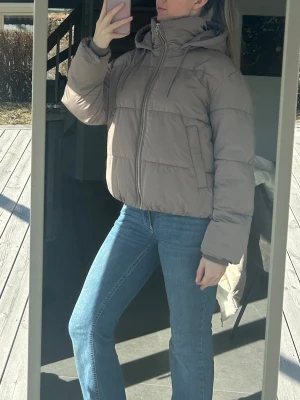 Brun pufferjacka med huva - Säljer en beige pufferjacka med dragkedja och huva. Jackan har en boxig passform, diskreta sidofickor och breda kanaler för extra värme. Perfekt för kalla dagar och enkel att styla till jeans eller andra byxor.