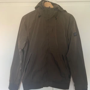 Olivgrön vindjacka från Weekend Offender - Snygg olivgrön vindjacka från Weekend Offender med huva som man kan gömma i kragen och dragkedja framtill. Jackan har två sidofickor med dragkedja och en bröst ficka och en diskret logotyp på ärmen. Perfekt för blåsiga dagar eller sommarn och enkel att matcha med olika outfits.