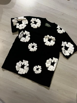 Svart t-shirt med vita blommor - Säljer en svart t-shirt från Denim Tears med stora vita blommor tryckta över hela tröjan. T-shirten har rund hals och korta ärmar, perfekt för dig som vill sticka ut med ett coolt och trendigt plagg.
