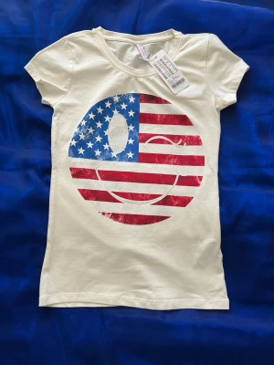 Vit t-shirt med amerikanskt flaggtryck - Snygg vit t-shirt med ett stort tryck av en amerikansk flagga i form av en cirkel på bröstet. T-shirten har rund hals och korta ärmar, perfekt för en avslappnad stil. Materialet känns mjukt och bekvämt, troligen bomull.