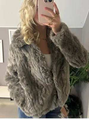 Grå fluffig pälsjacka - Säljer en supermjuk och fluffig grå pälsjacka med bred krage och avslappnad passform. Jackan har långa ärmar och är perfekt för kalla vinterdagar. Materialet är fuskpäls som ger en lyxig känsla och trendig look.