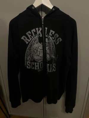 Reckless Scholars hoodie  - Svart hoodie från Reckless Scholars med dragkedja framtill och huva. några få stenar saknas på trycket men inget man tänker på.