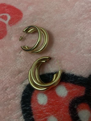 Guldiga chunky hoops örhängen - Säljer ett par trendiga chunky hoops örhängen i guldfärgad metall. De har en dubbelslingad design som ger ett coolt och modernt intryck. Perfekta för att lyfta vilken outfit som helst med en touch av guld. En plopp saknas för att stänga den