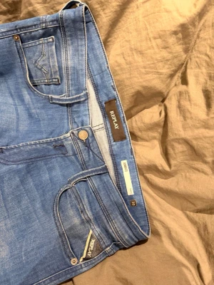 Blå slim fit jeans från Replay - Snygga blå slim fit jeans från Replay med klassisk femficksdesign och ljusa slitningar på låren. Jeansen har en modern passform och detaljerade sömmar, samt Replay-logga på fickan. Perfekta för en trendig och avslappnad stil. Inte använda på ett bra tag då de har varit för små nypris är oklart:)