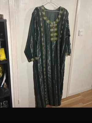 Grön långklänning Xl/L - Unik grön långklänning med vertikala ränder i silver och dekorativa guldiga detaljer vid halsringning och ärmslut. Klänningen har lös passform och trekvartsärm, perfekt för dig som vill ha något bekvämt och stilrent.