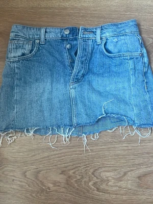 Blå jeanskjol - Säljer en kort blå jeanskjol från Zara med fransig nederkant och klassiska fem fickor. Kjolen har dubbla knappar framtill och bälteshällor. Perfekt för dig som gillar en avslappnad och trendig stil.