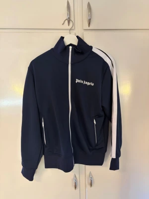 Palm Angels marinblå ziptröja - Snygg marinblå ziptröja från Palm Angels med vit dragkedja och vita detaljer på ärmarna. Tröjan har hög krage, två fickor med dragkedja och logga på bröstet. Perfekt för en sportig och trendig look.